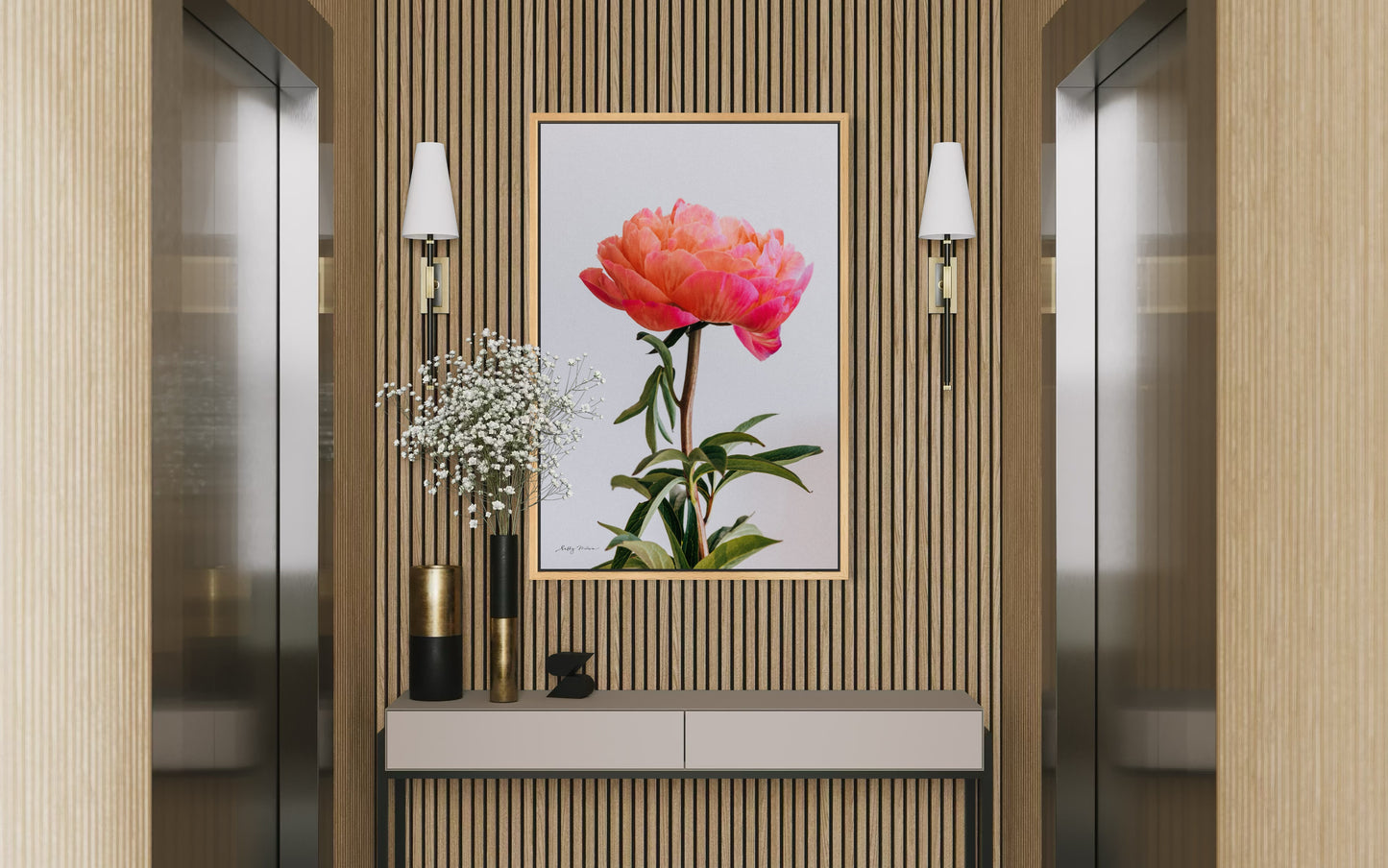 A fine art photography print of a bright pink peony set on a white background. The print is framed in natural wood colour and set on a wall between two elevators in an office building.