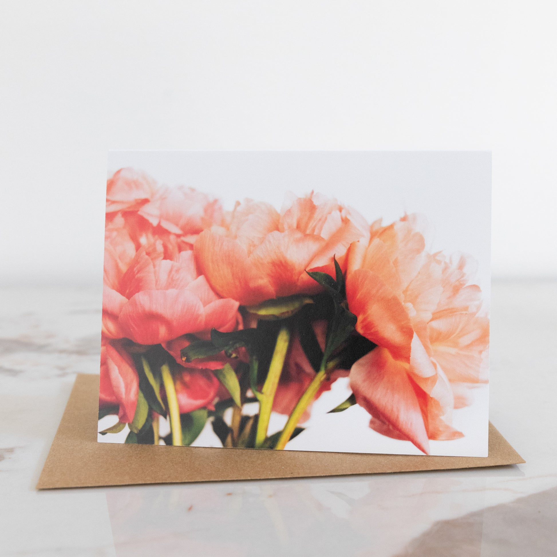 A peony flower fine art photography print greeting card, sat up on a marble and white background. It has a brown envelope. The peonies displayed on the card are in a bunch.  It is in landscape aspect.