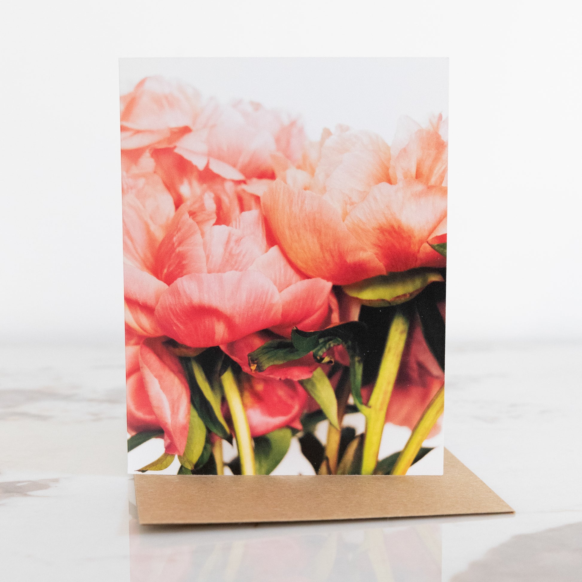 A peony flower fine art photography print greeting card, sat up on a marble and white background. It has a brown envelope. The peonies displayed on the card are in a bunch.  This is in a portrait aspect. 