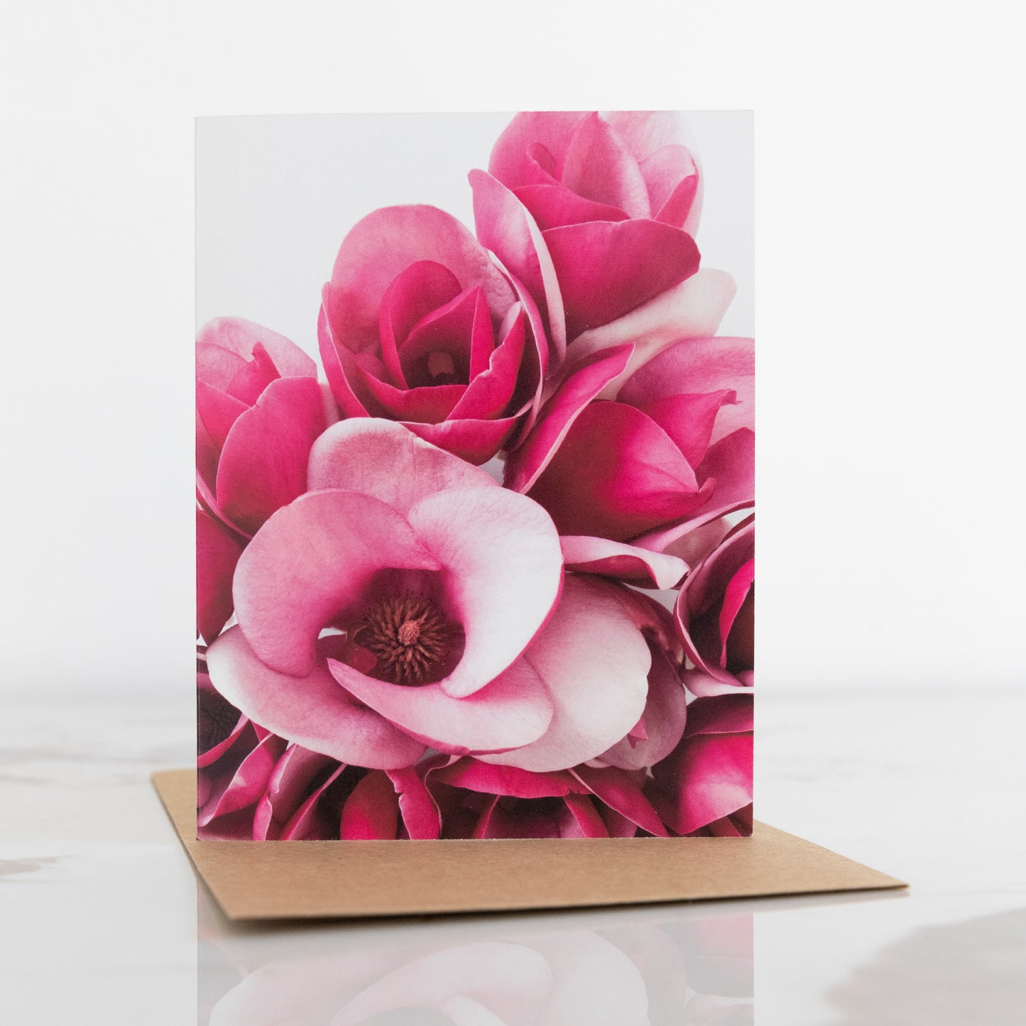 A magnolia flower fine art photography print greeting card, sat up on a marble and white background. It has a brown envelope. This is a print in portrait aspect